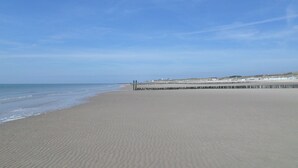 Una extensa playa arenosa con olas suaves, cielo despejado y un muelle distante.