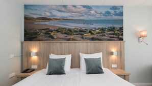 Un dormitorio con cabecera de madera, ropa de cama blanca, dos almohadas, lámparas de noche y un cuadro con una playa.