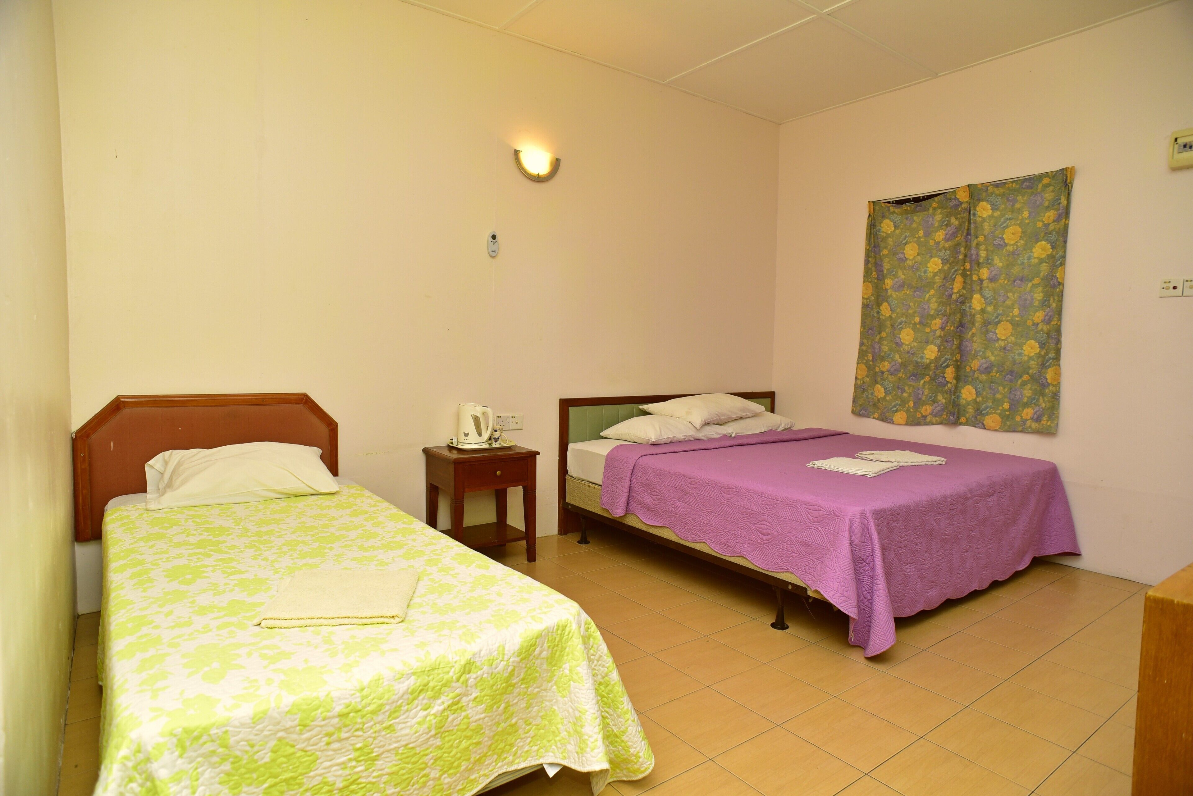 A room with two beds, a nightstand, a wall-mounted light, and a floral-patterned curtain.