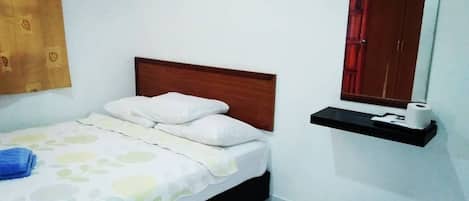 A neatly made bed with a wooden headboard, a small shelf with a mirror and a roll of toilet paper, and a patterned bedspread.