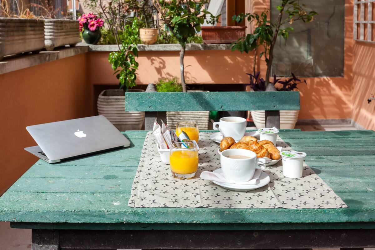 A table set with breakfast items including croissants, coffee, and juice, with a laptop and newspapers on a green tablecloth.
