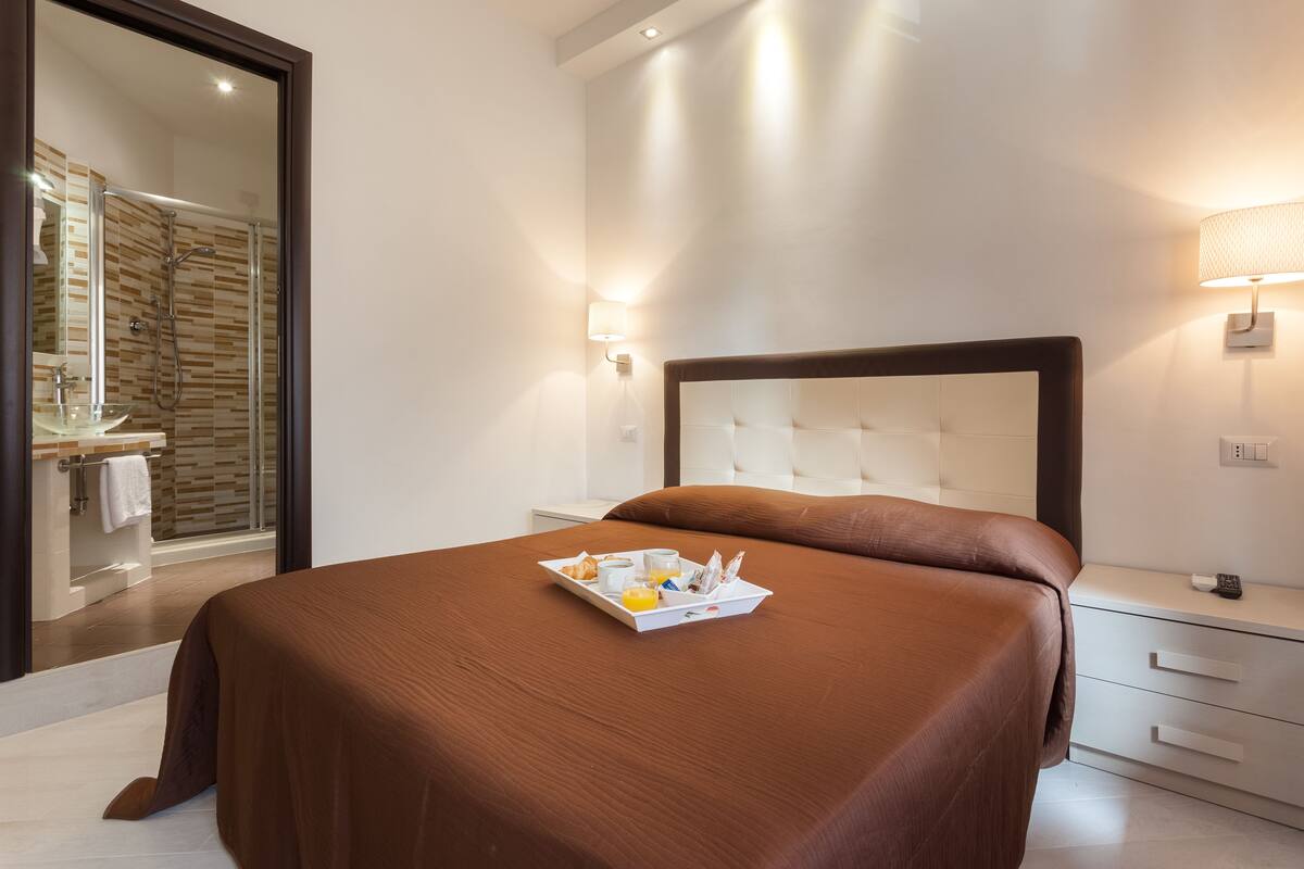 A modern hotel room with a large bed, a bedside table, a nightstand, a glass shower enclosure, and a bathroom visible through an open door.