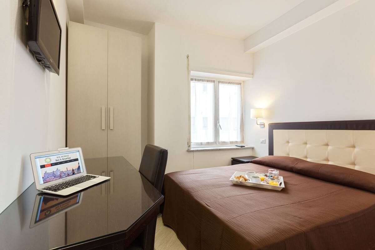 A hotel room with a bed, a desk with a laptop, a bedside table with a tray of breakfast items, and a window with curtains.