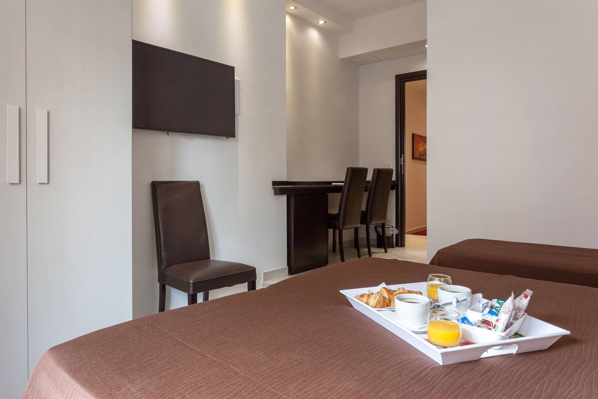 A hotel room with a bed, a breakfast tray with croissants, coffee, and orange juice, and a TV mounted on the wall.