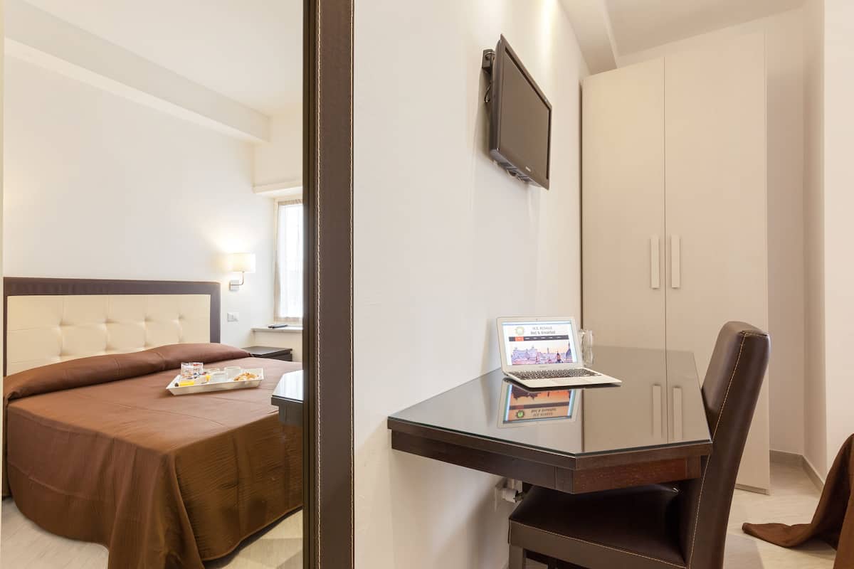A hotel room with a bed, a desk with a laptop, a TV on the wall, and a wardrobe.