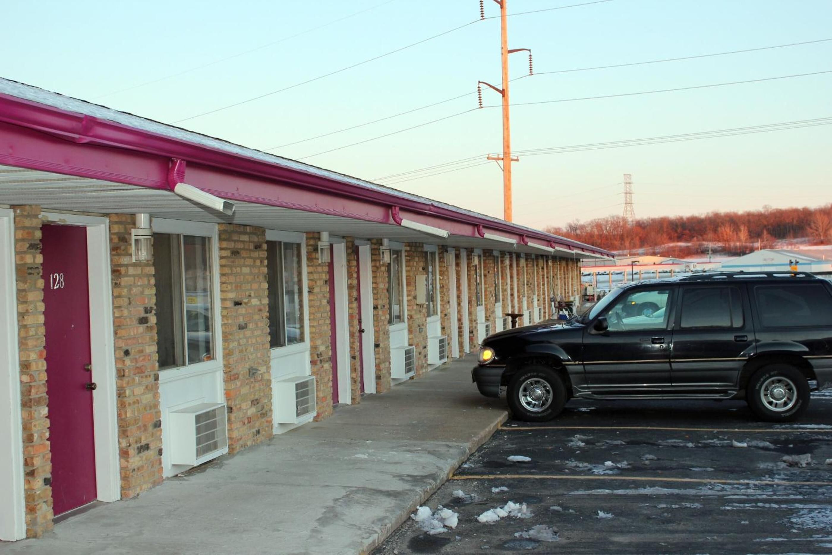 A motel with a row of rooms, each with a door and window, and a parking lot with a black SUV parked in front.