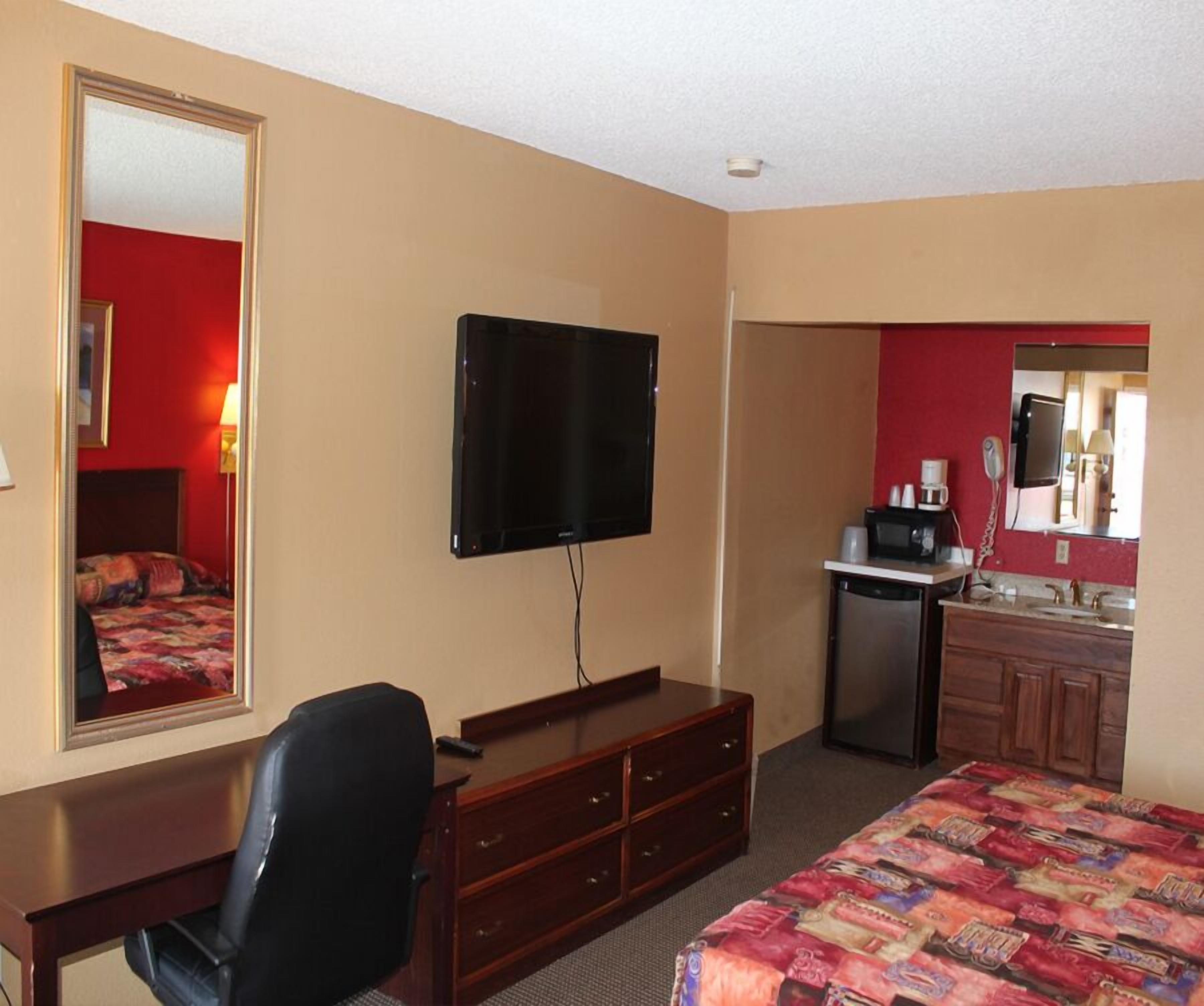 A hotel room with a bed, a desk, a chair, a television, a dresser, a mirror, a coffee maker, and a refrigerator.