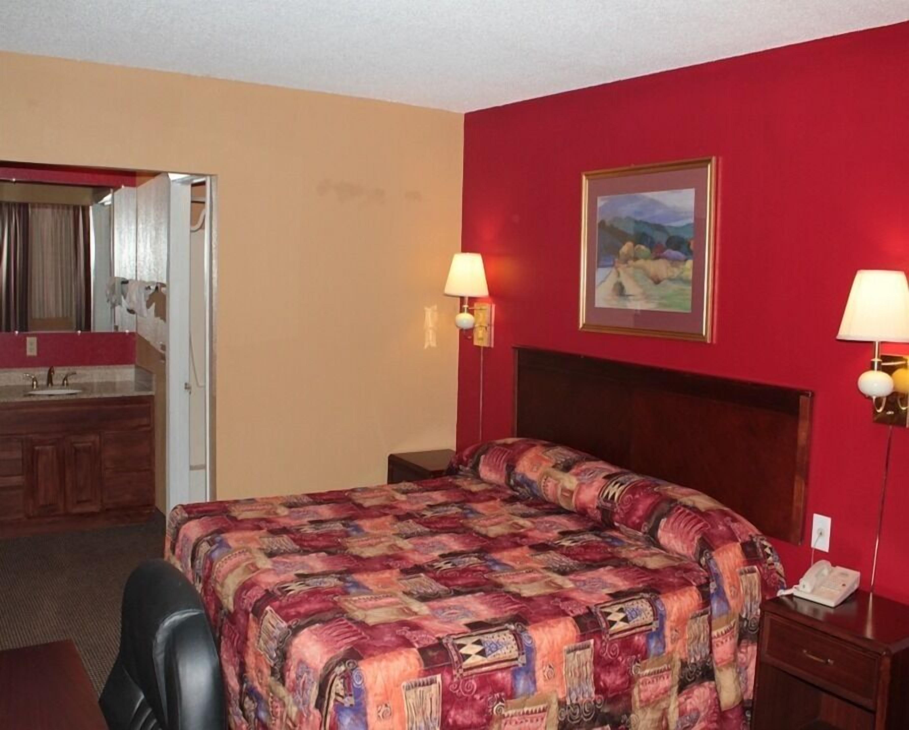 Habitación de hotel con una cama grande, cabecera de madera, un cuadro en la pared, dos lámparas de noche, un teléfono y un baño visible a través de una puerta abierta.