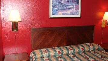 Una habitación de hotel con cabecera de madera, cama con colcha estampada de flores, mesita de noche con lámpara y un cuadro enmarcado en la pared.