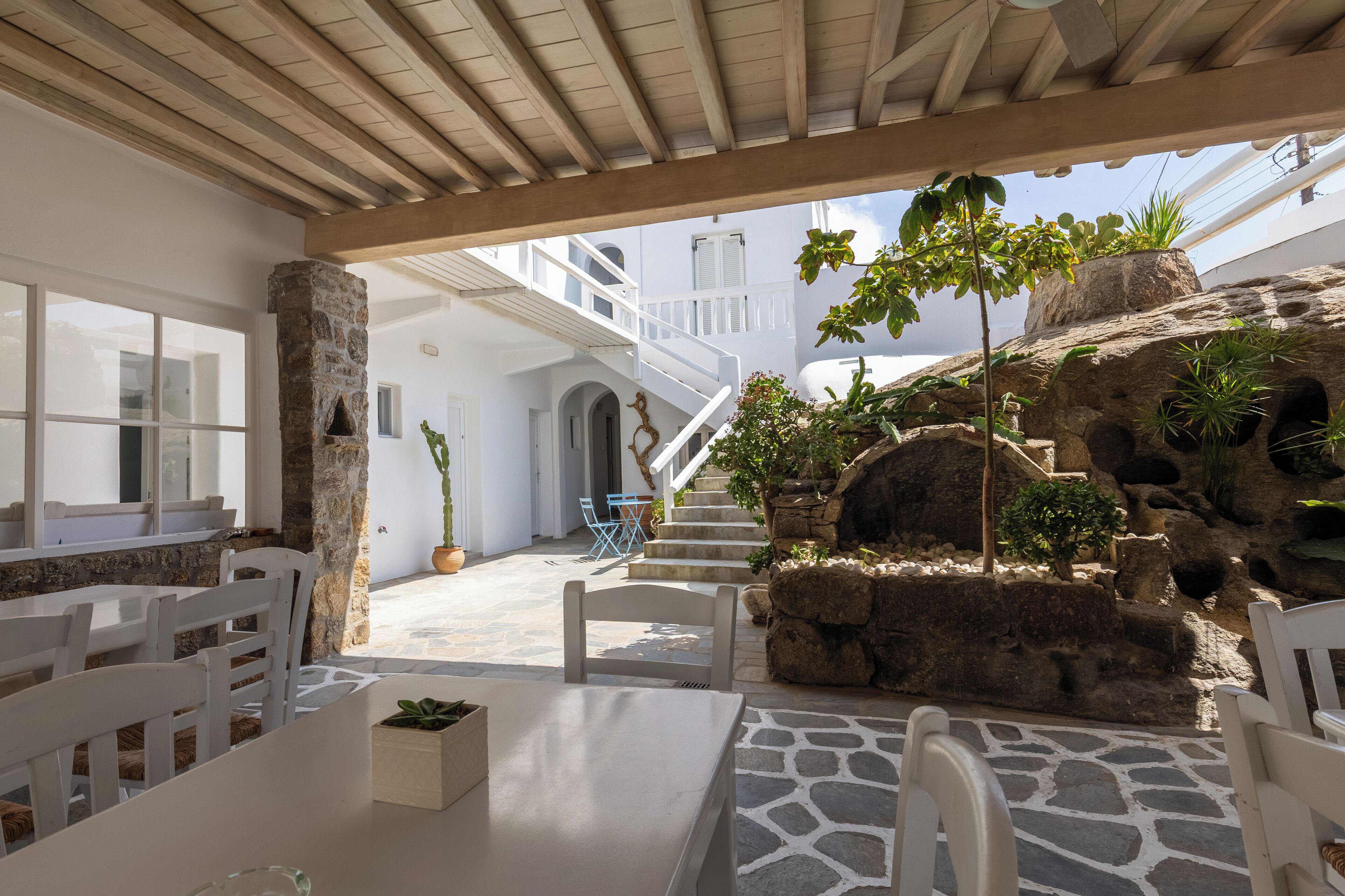 A covered outdoor seating area with stone flooring, white furniture, and a view of a white building with a staircase.