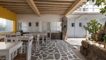 Un área de comedor al aire libre con mesas y sillas blancas, piso de piedra y techo de madera.
