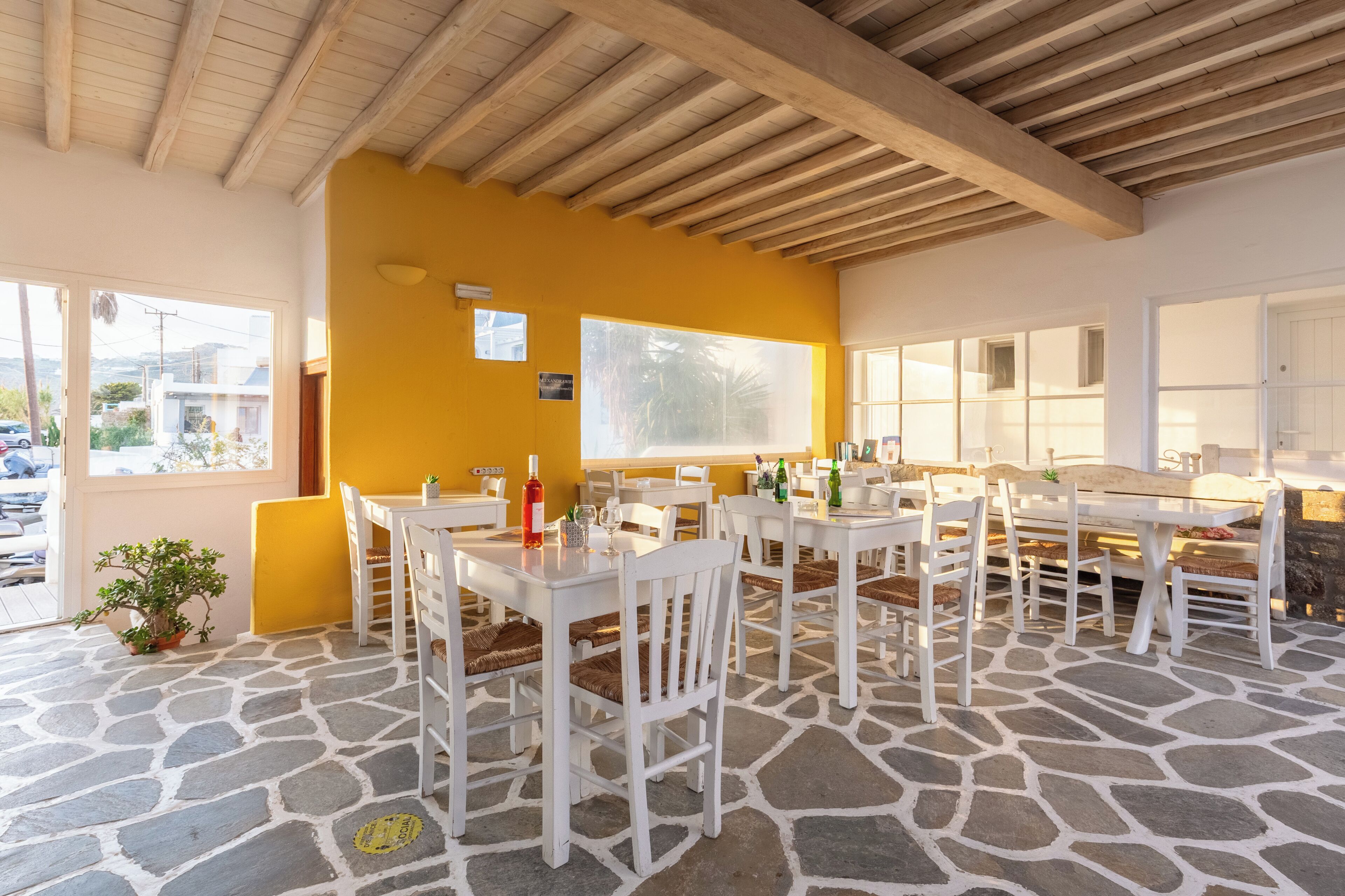 Un espace extérieur aménagé pour les repas, avec des tables et des chaises blanches, un mur jaune et un sol en pierre.