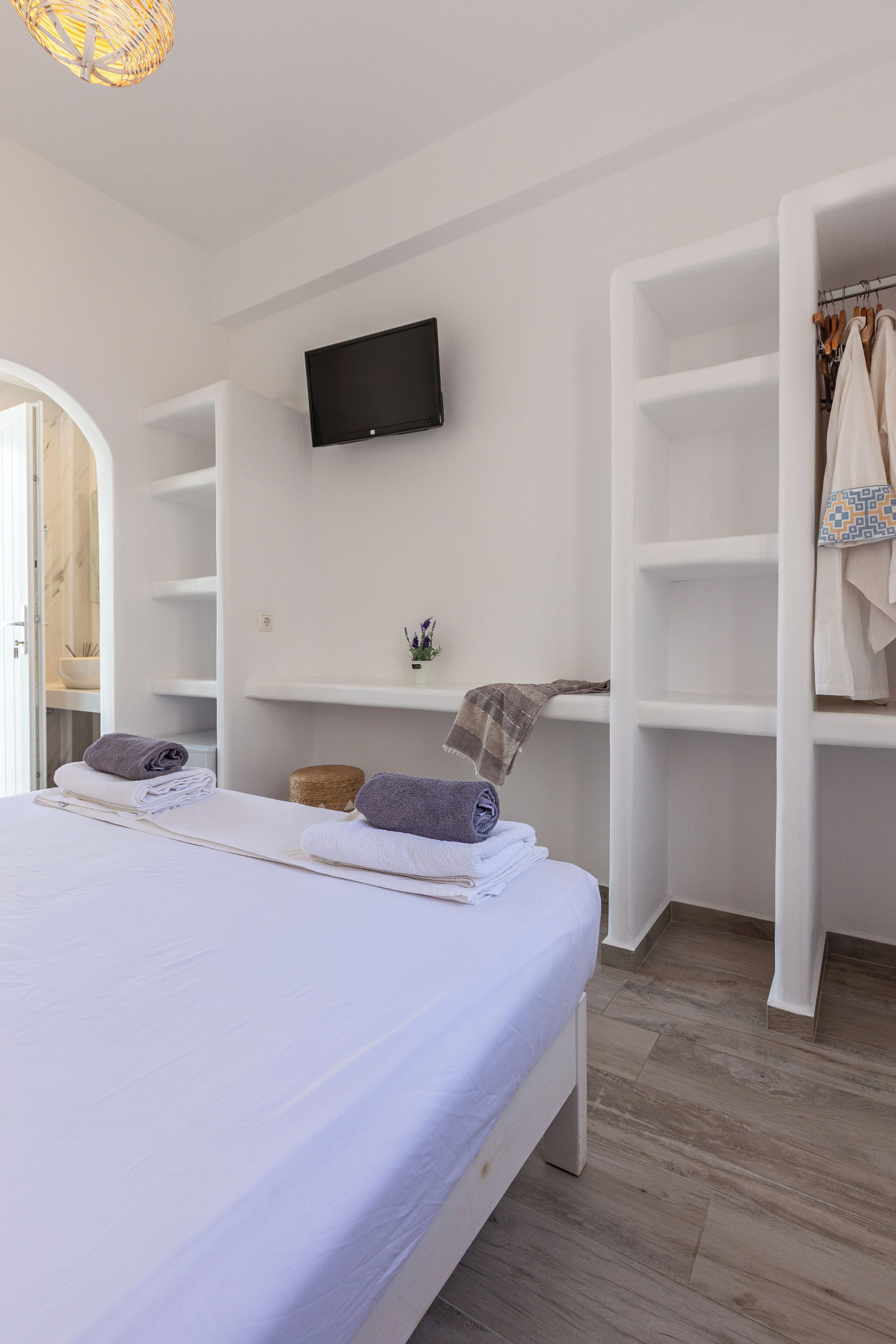 A bedroom with a bed, a flat-screen TV mounted on the wall, and a wardrobe with hanging clothes.
