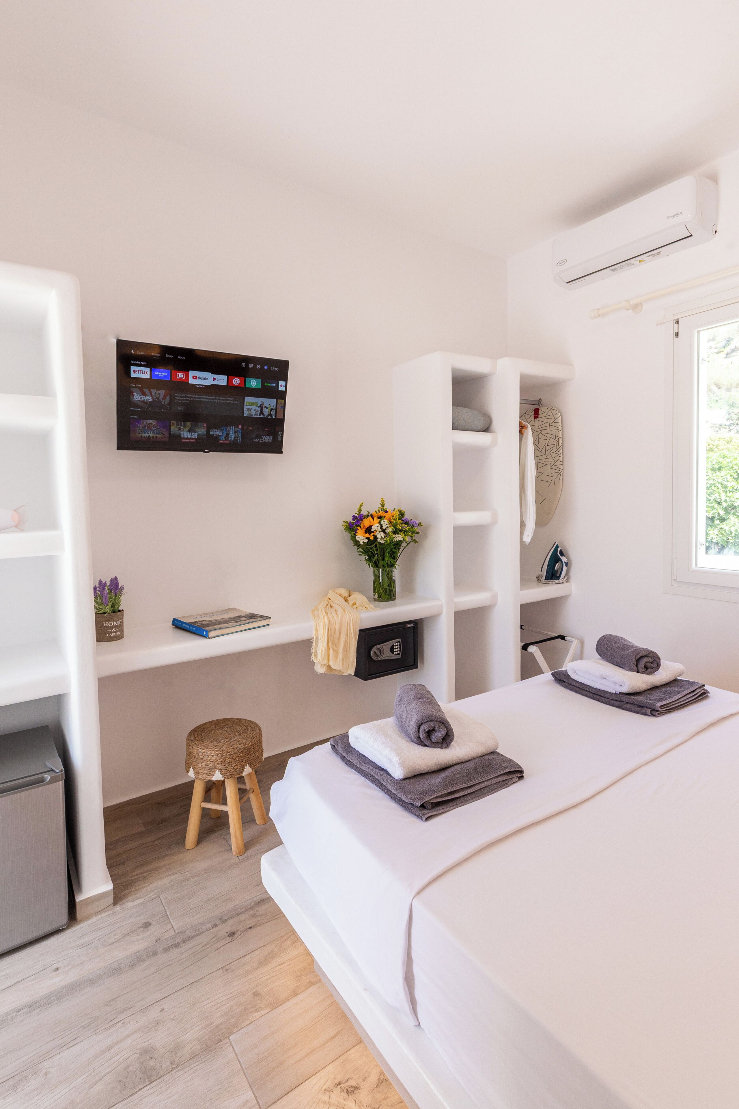A modern bedroom with a bed, a TV mounted on the wall, a small stool, and a shelf with a vase of flowers.