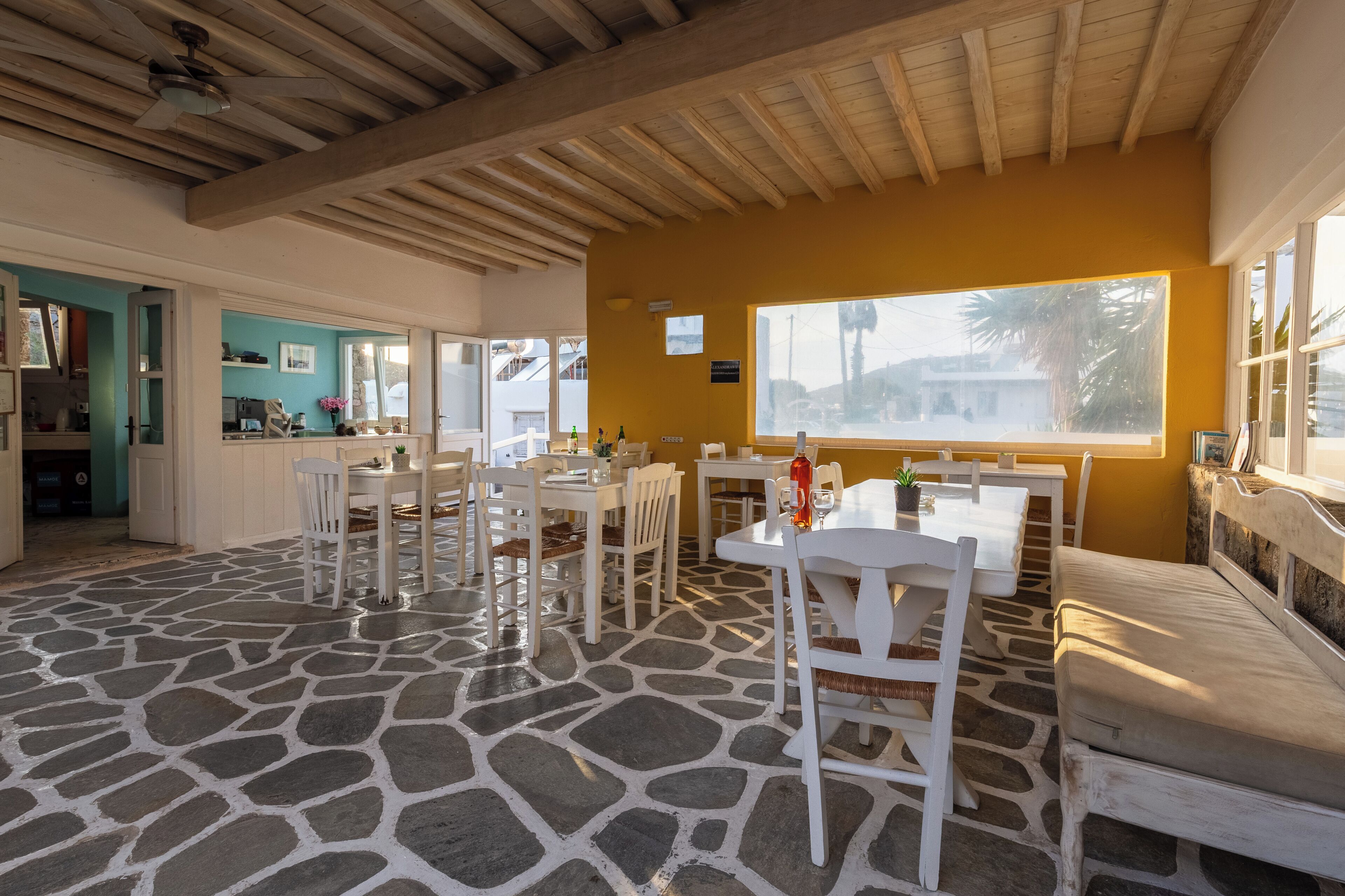 A dining area with white tables and chairs, a stone floor, and a view through a large window.