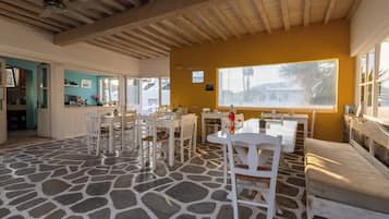 A dining area with white tables and chairs, a stone floor, and a view through a large window.