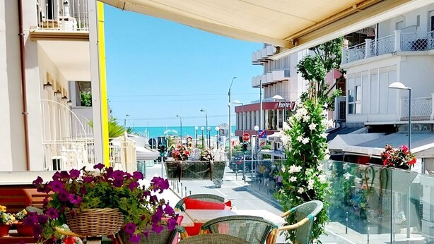 An outdoor seating area with wicker chairs and tables, a wicker basket with flowers, and a view of the sea and buildings in the background. - Hotel Brotas (Rimini)