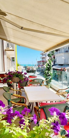 An outdoor seating area with wicker chairs and tables, a wicker basket with flowers, and a view of the sea and buildings in the background.