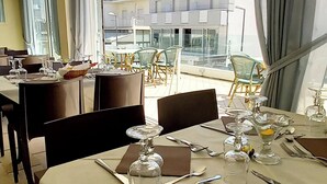 A dining area with tables set for two, large windows with curtains, and a view of a modern building with balconies. - Hotel Brotas (Rimini)
