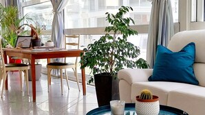 A modern hotel room with a large window, a dining area with a wooden table and chairs, a sofa with a blue pillow, and a coffee table with a cactus and starfish decoration. - Hotel Brotas (Rimini)