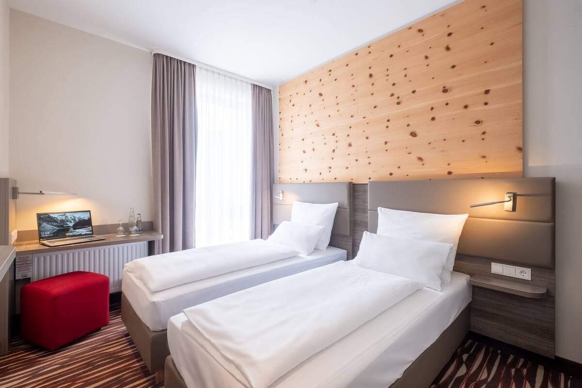 A hotel room with two beds, a desk with a laptop, a red ottoman, and a wooden accent wall.