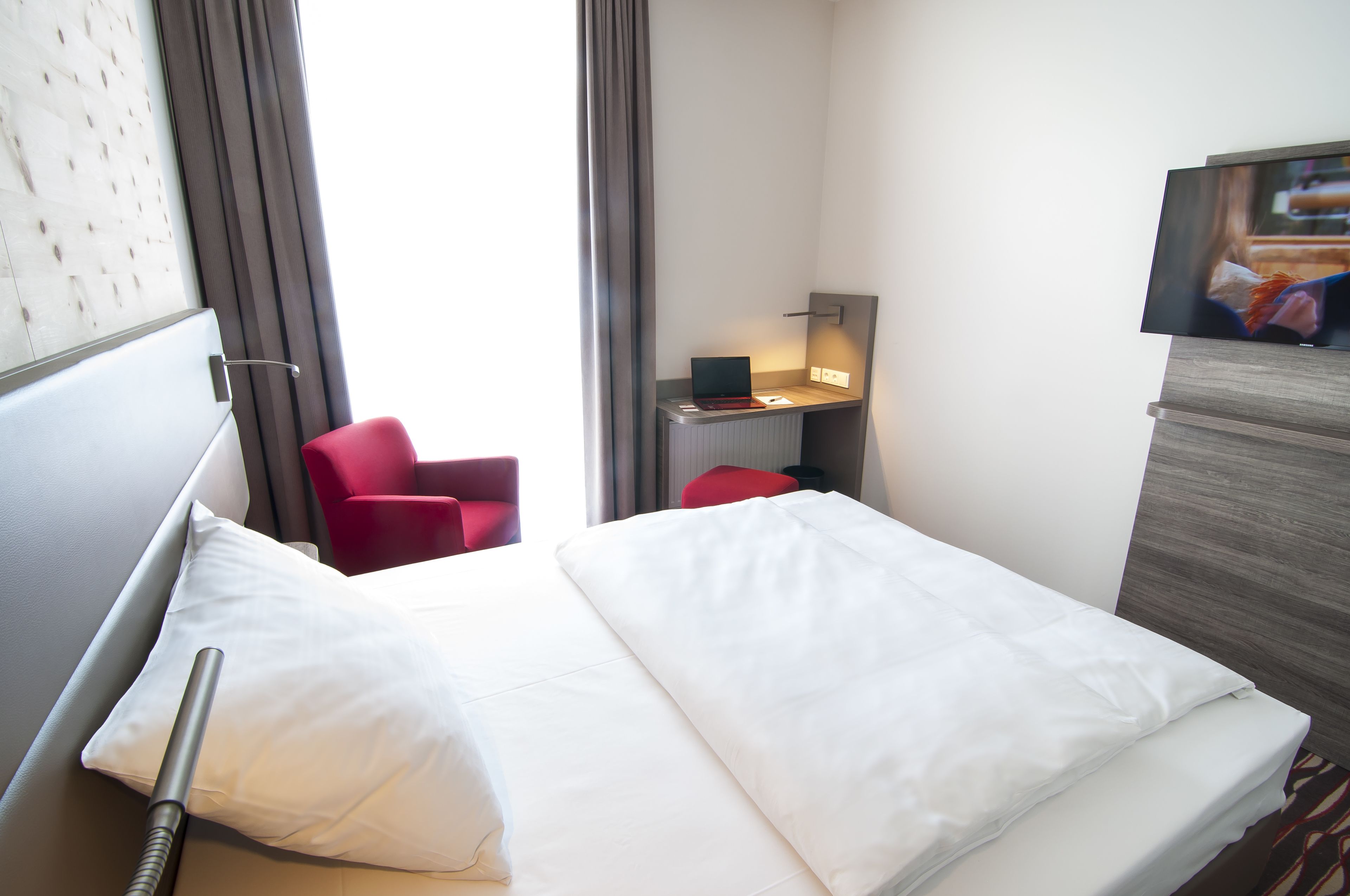 A hotel room with a bed, a desk with a laptop, a red chair, and a TV on a wooden cabinet.