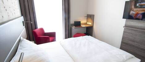 A hotel room with a bed, a desk with a laptop, a red chair, and a TV on a wooden cabinet.