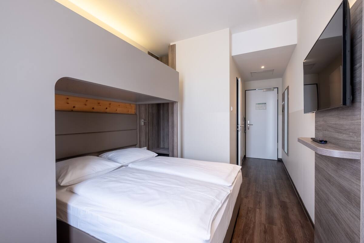 A modern hotel room with a large bed, a wooden headboard, a flat-screen TV mounted on the wall, and a wooden floor.