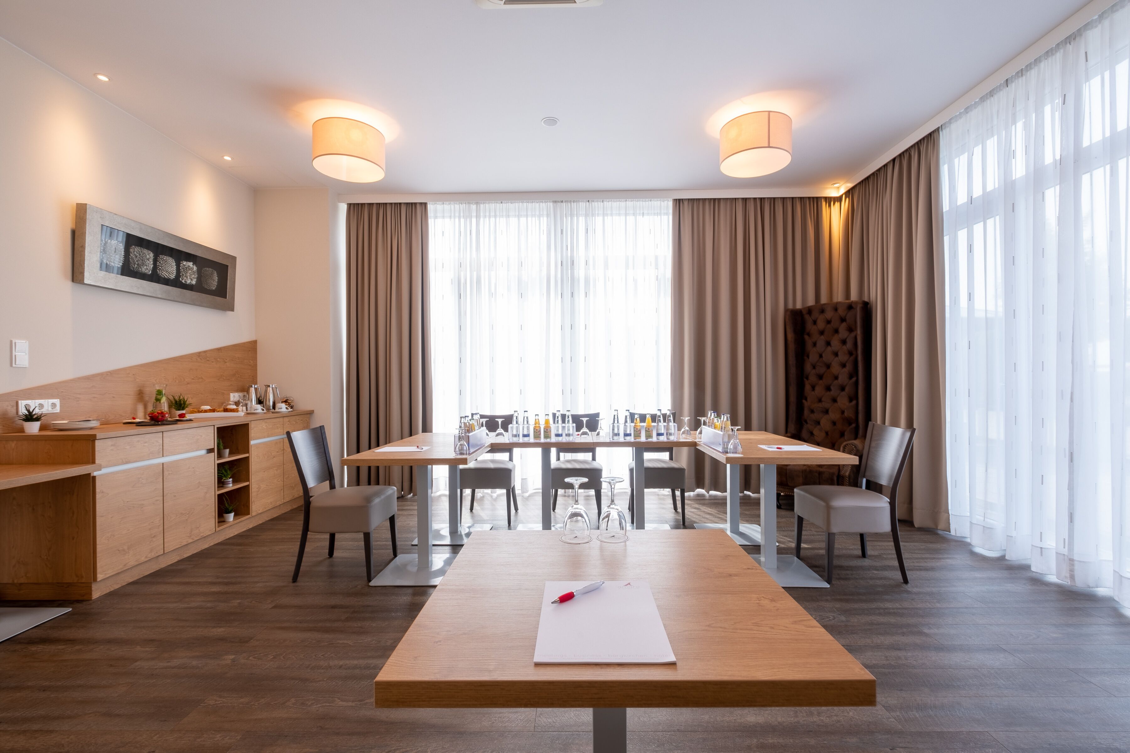 Ett modernt hotellrum med en matplats, möbler i trä och stora fönster med gardiner.