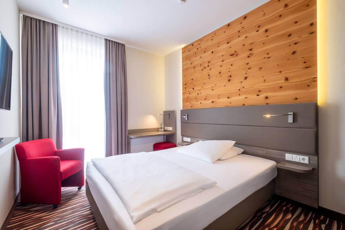 A hotel room with a large bed, a red armchair, a desk with a lamp, and a wooden accent wall.