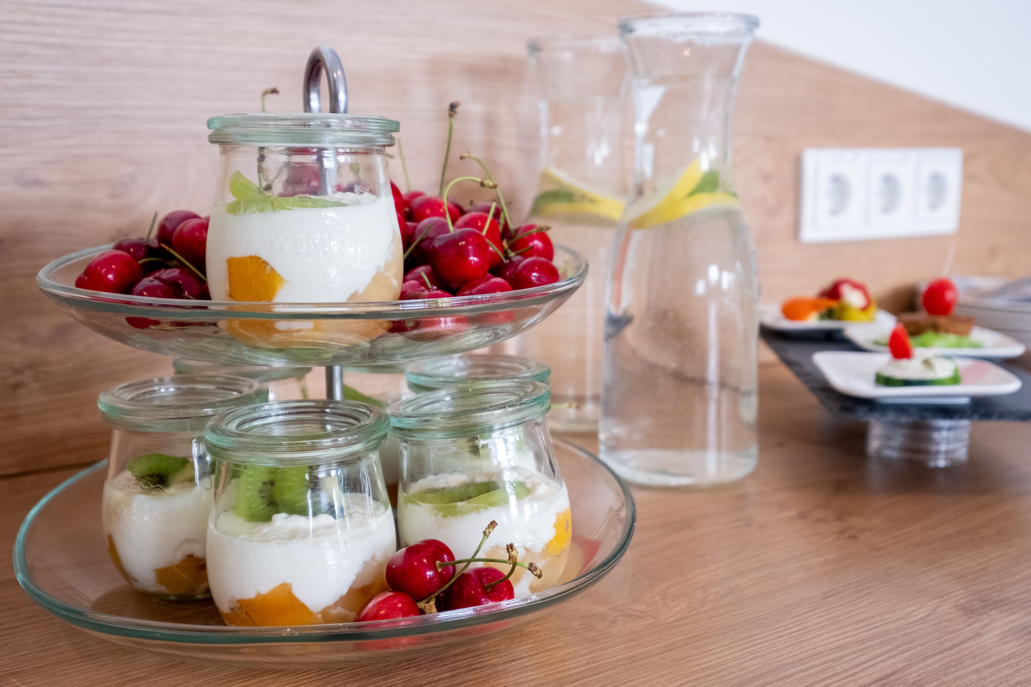 Un elegante expositor de vidrio de tres niveles con parfait de fruta y yogur, una jarra de vidrio transparente y un plato pequeño con aperitivos.