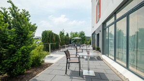 Un patio al aire libre con mesas y sillas, rodeado de follaje y una fachada de edificio moderna.