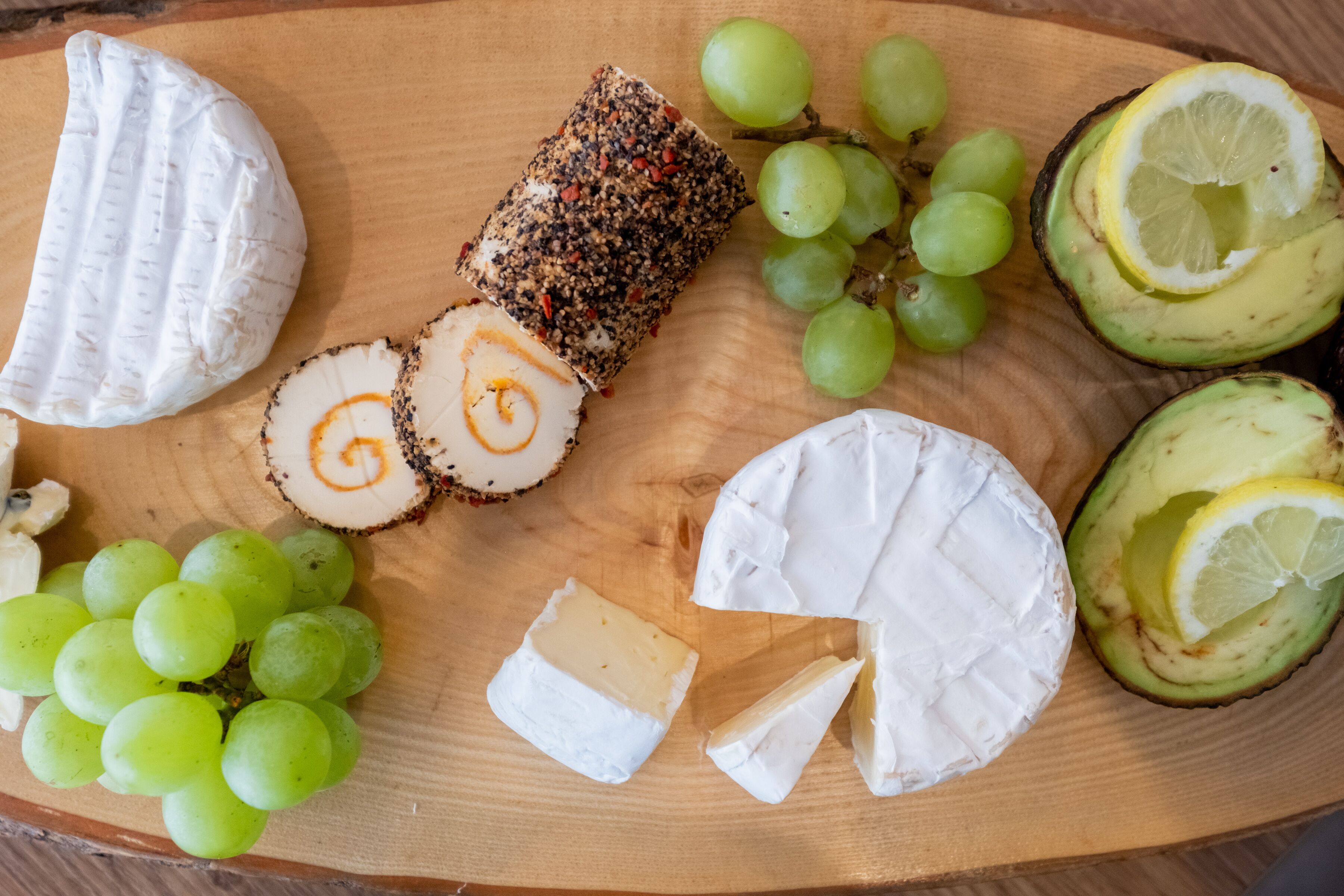 A wooden board with various cheeses, grapes, and avocado slices.