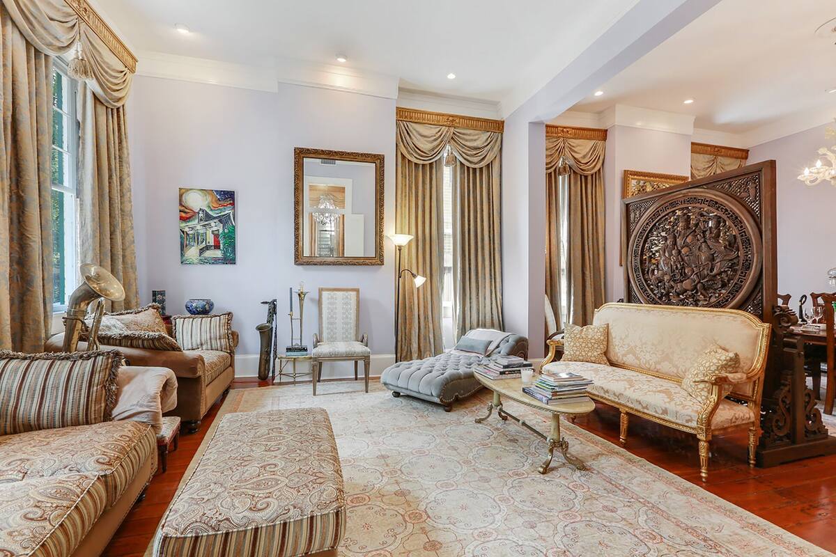 A spacious living room with a large sofa, a smaller armchair, a coffee table, and a patterned rug. There are two large windows with curtains, a chandelier, and a framed artwork on the wall.