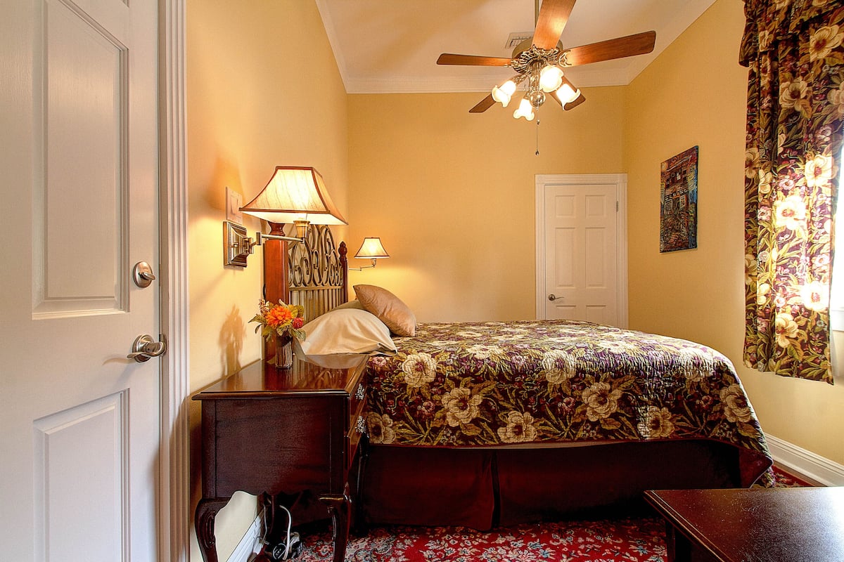 A bedroom with a floral bedspread, a ceiling fan, a nightstand with lamps, and a door.