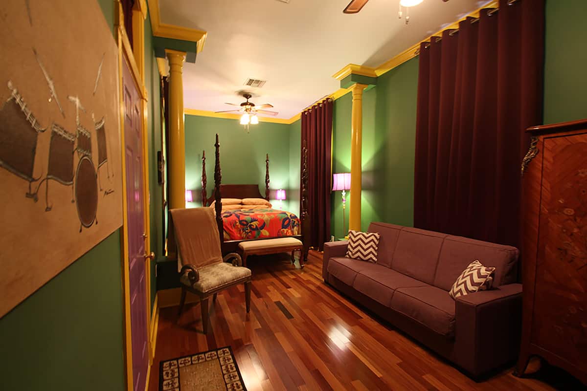 A room with a wooden floor, a bed with a colorful blanket, a purple sofa, a chair, and a ceiling fan.