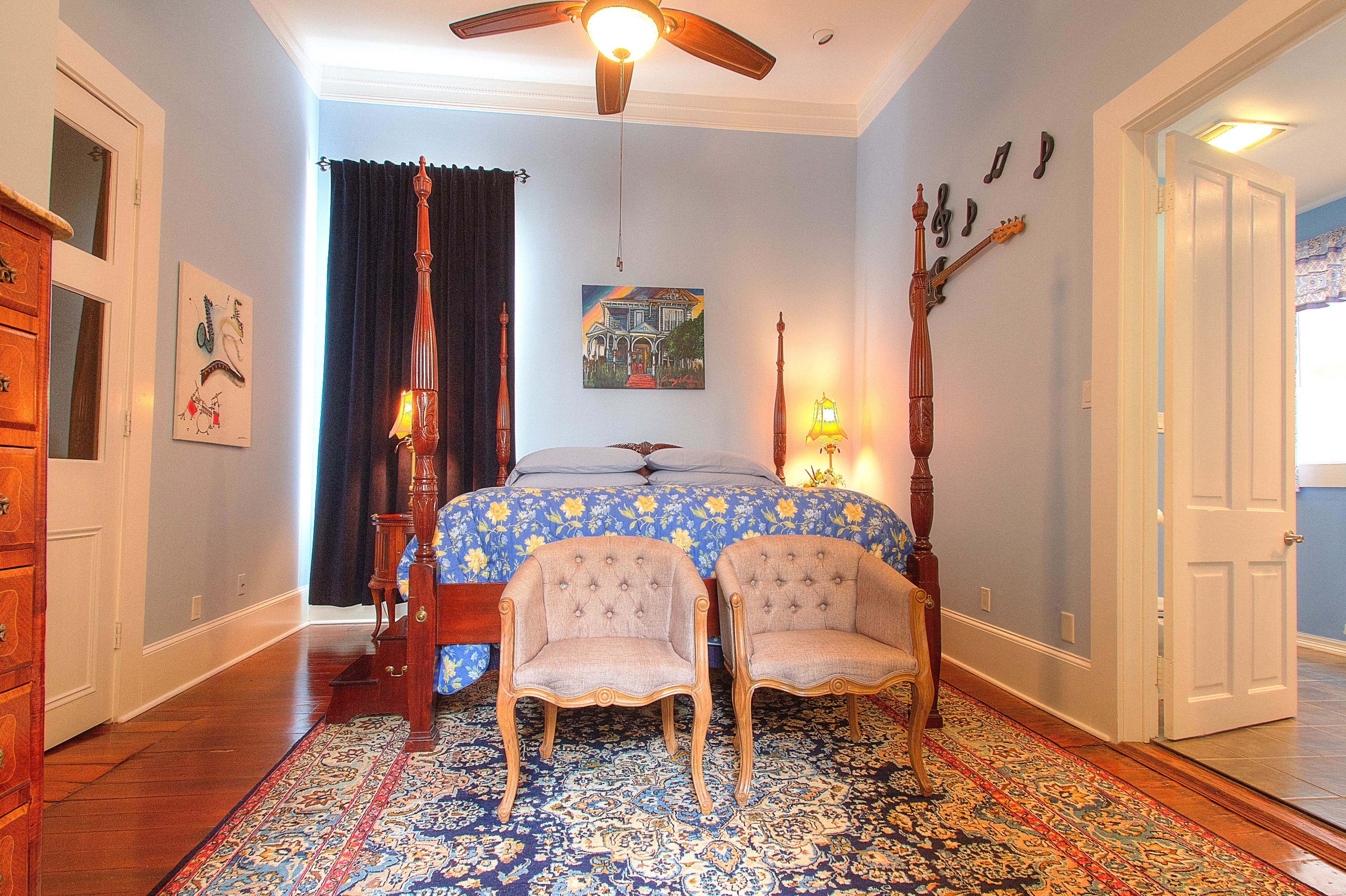 A bedroom with a bed, two chairs, a ceiling fan, a rug, and a painting on the wall.