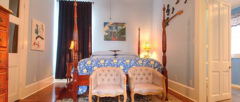 A bedroom with a bed, two chairs, a ceiling fan, a rug, and a painting on the wall.