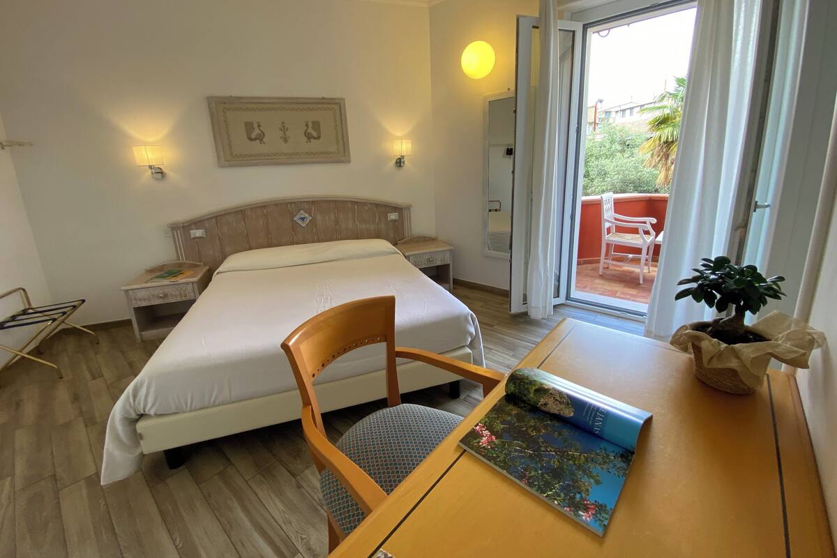 A hotel room with a bed, a wooden chair, a table with a book, a nightstand, a wall-mounted lamp, and a sliding glass door leading to an outdoor area.