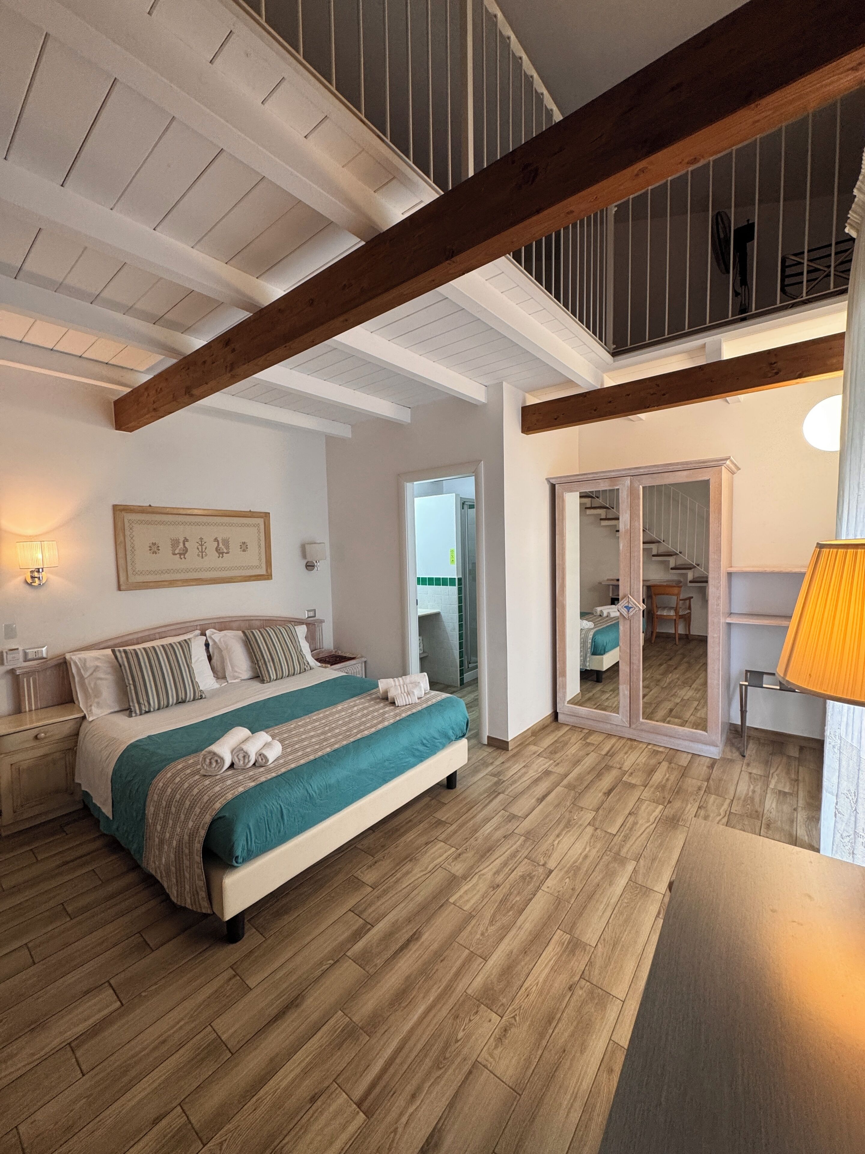 Une chambre à coucher avec un lit, un plancher en bois, un plafond avec des poutres apparentes, une porte donnant sur une salle de bain et une lampe sur une table de chevet.