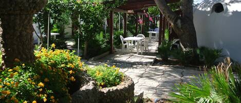 Un patio jardín con suelo de piedra, mesas y sillas blancas bajo una pérgola.