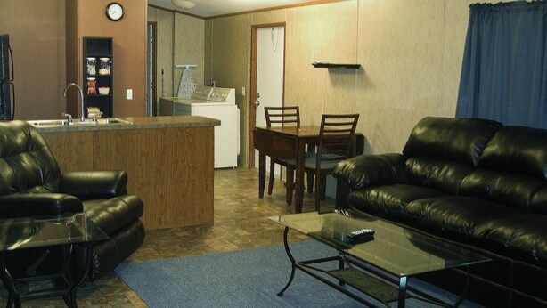 A living room with a leather sofa, a coffee table, a TV stand, and a kitchen area in the background. - Eagle Ford Lodging (Pleasanton)