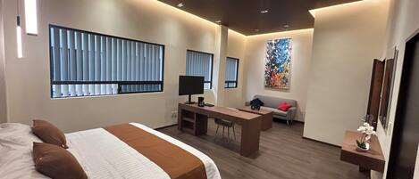 A modern hotel room with a large bed, a desk, a sofa, and a large window with blinds.