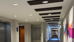Un couloir d’hôtel moderne, avec un tracé rectiligne, des ascenseurs et une œuvre d’art murale abstraite aux couleurs vives.