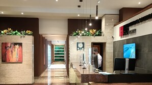Un hall d’hôtel moderne, doté d’un comptoir d’accueil, d’un écran numérique et d’un long couloir menant à un autre espace.