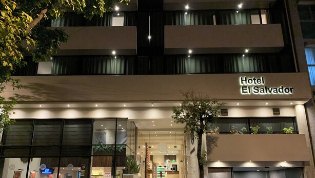 L’entrée d’un hôtel, avec une enseigne indiquant « Hôtel El Salvador ». Des marches mènent à l’entrée, un petit arbre se trouve sur la droite, et une personne marche sur le trottoir.