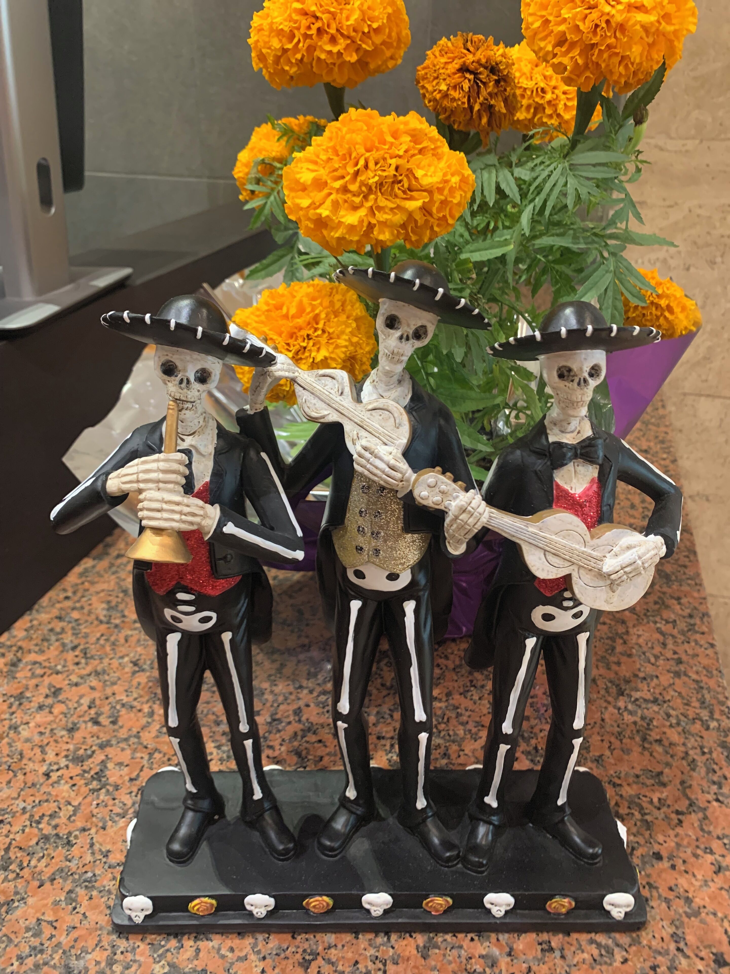 Uma pequena escultura decorativa de esqueleto sobre uma bancada de granito, com três esqueletos vestidos de preto e detalhes brancos, segurando instrumentos musicais e usando sombreros.