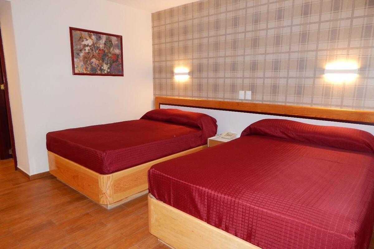 Une chambre avec deux lits en bois, chacun recouvert d’une courtepointe rouge, doté d’une tête de lit à carreaux et orné d’un tableau au mur.