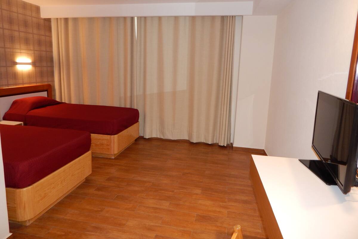 Une chambre d’hôtel avec deux lits simples, une télévision et une grande fenêtre avec des rideaux.