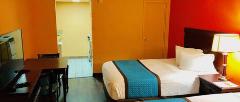 A hotel room with two beds, a desk, a chair, a TV, and a lamp.